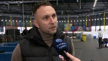 Mężczyzna w kamizelce i golfie udziela wywiadu, trzymając mikrofon z logo "Polsat Sport". W tle widoczna jest lodowa hala sportowa z kibicami i zawodnikami.