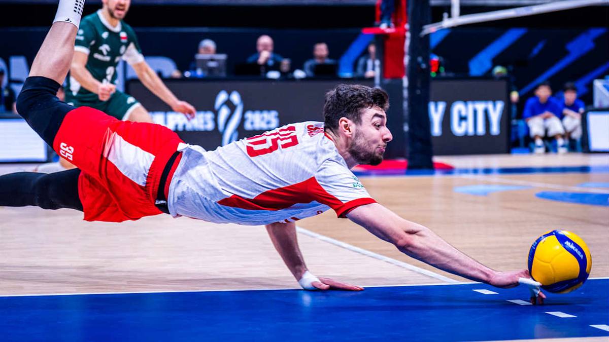 Siatkarz w białej koszulce z czerwonymi wstawkami i czerwonych spodenkach, wykonuje obronę na boisku do siatkówki, próbując odbić piłkę.