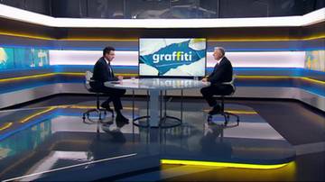 Dw&oacute;ch mężczyzn w garniturach siedzi przy okrągłym stole w studiu telewizyjnym, przed nimi duży ekran z napisem "graffiti".