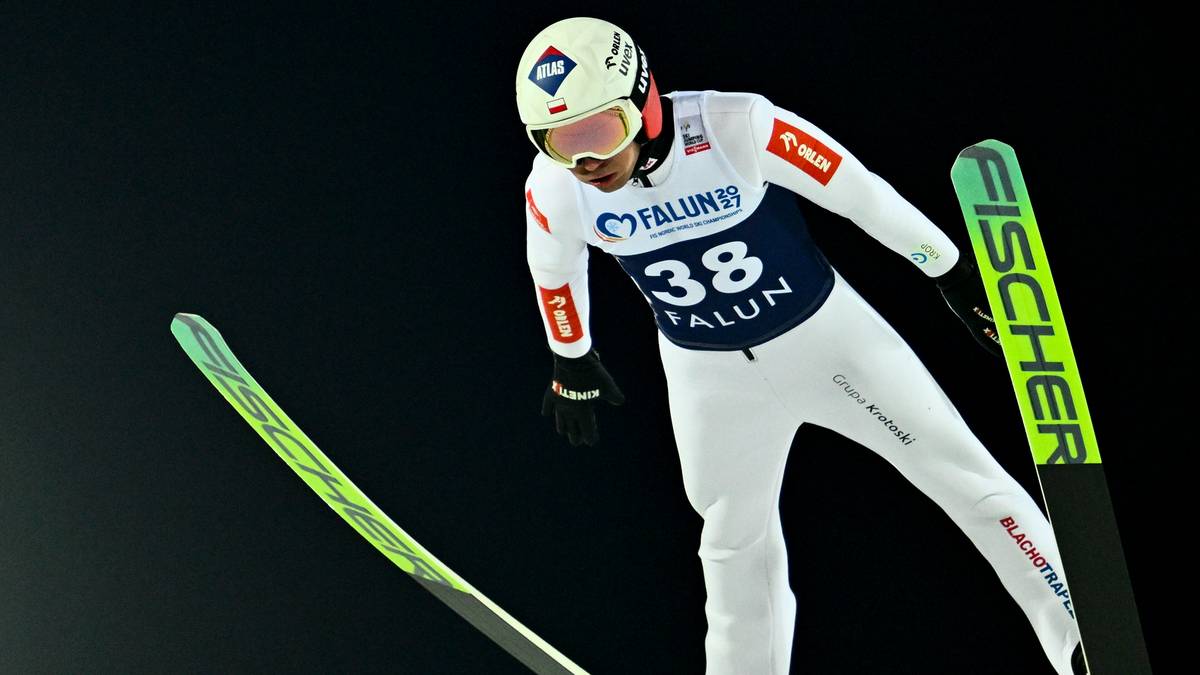 Skoczek narciarski w białym stroju z numerem 38 na kombinezonie, stojący na skoczni narciarskiej.