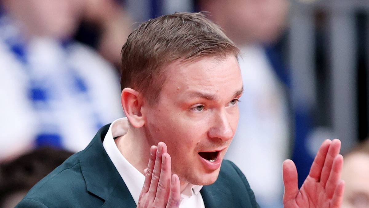 Trener w garniturze i białej koszuli, z otwartymi dłońmi i wyrazistą miną, podczas meczu koszykówki.