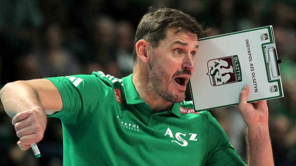 Mężczyzna w zielonej koszulce z napisem "ASZ" trzyma planszę taktyczną z logo "Indykpol AZS Olsztyn". Jego usta są szeroko otwarte, a wyraz twarzy sugeruje zaangażowanie i emocje.