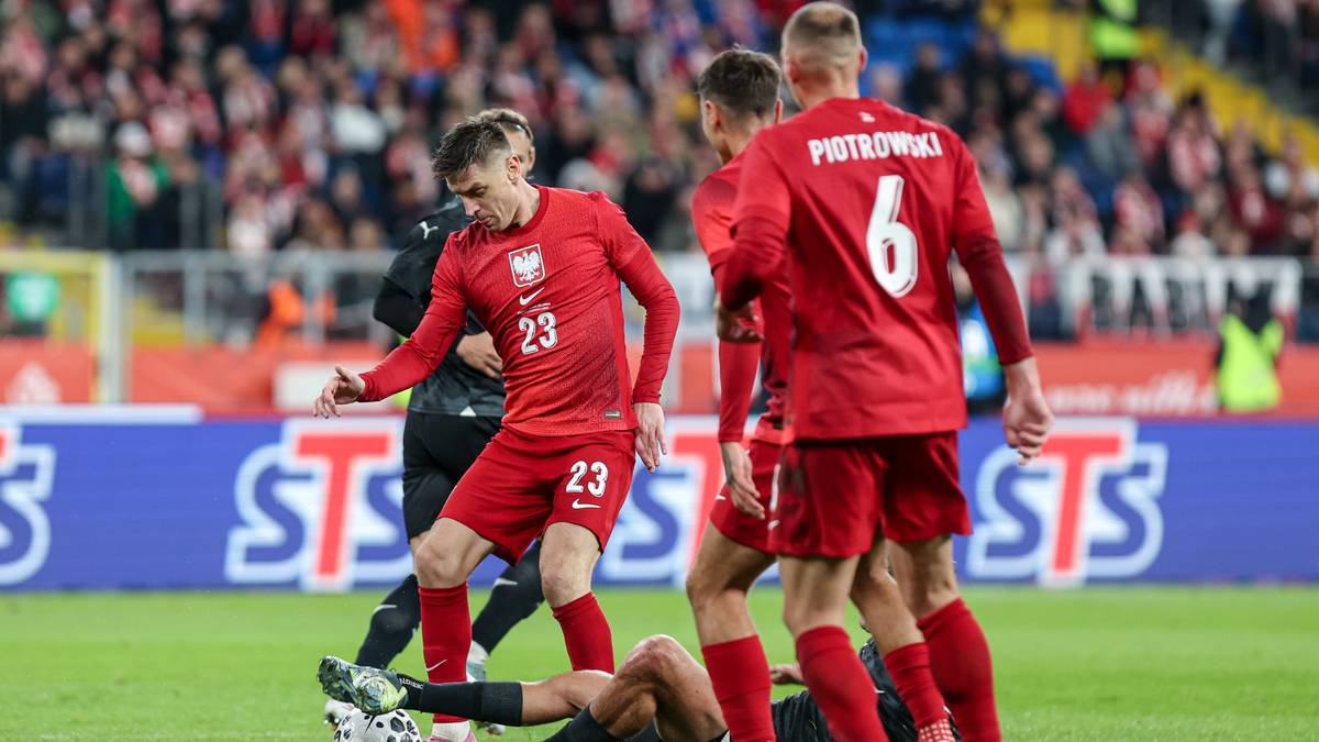 Piłkarze w czerwonych strojach narodowych ze stopami na boisku, jeden z nich leży na ziemi, inny stoi nad nim.