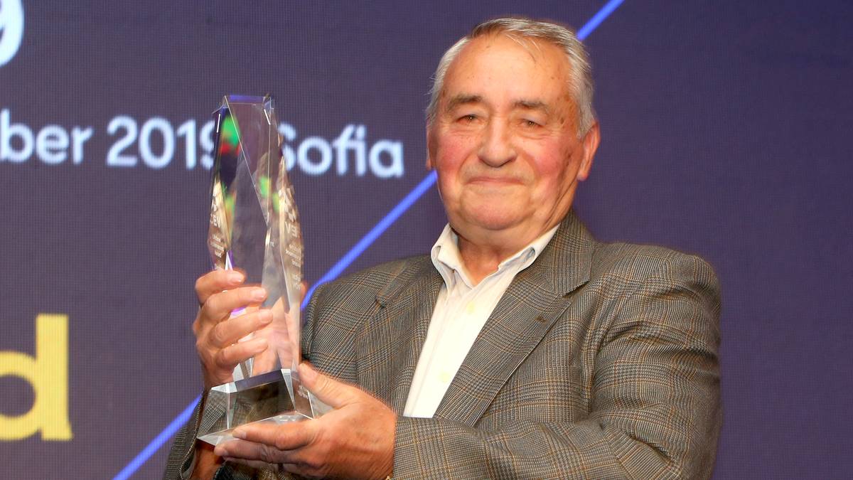 Starszy mężczyzna w szarym garniturze i białej koszuli, z uśmiechem na twarzy, trzyma w rękach dużą, przezroczystą statuetkę. W tle widać fragment napisu "ber 2019 Sofia" oraz niebieskie linie tworzące geometryczny wzór.