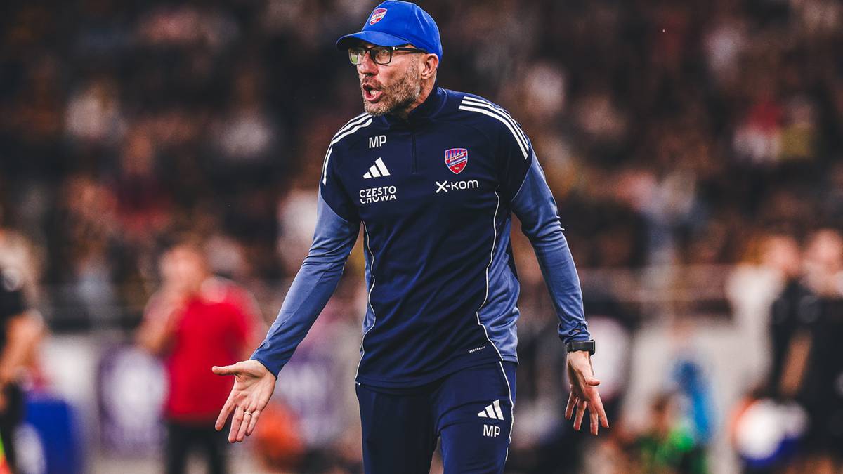 Trener piłkarski w niebieskiej czapce i okularach, ubrany w granatową bluzę sportową z niebieskimi rękawami i białymi napisami, stoi na boisku z wyciągniętymi dłońmi i wyrazem twarzy sugerującym frustrację.