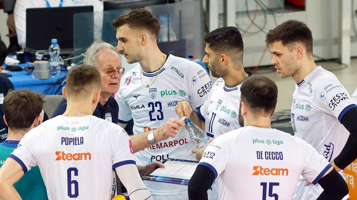 Drużyna siatkarzy w białych strojach stoi w kręgu na boisku, omawiając taktykę. Zawodnicy mają numerki na koszulkach, jeden z nich trzyma piłkę.