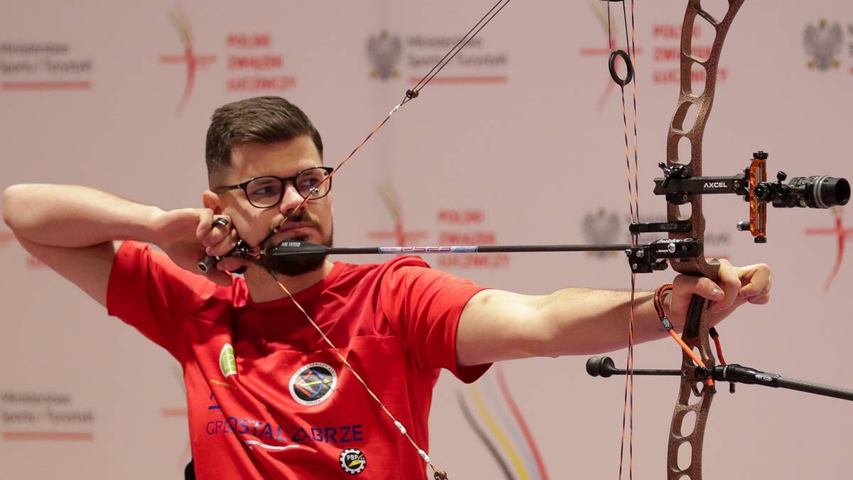 Mężczyzna w czerwonej koszulce i okularach celuje z łuku do tarczy podczas zawodów.