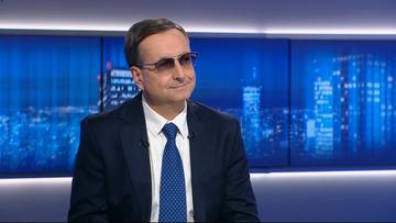 Marek Magierowski w okularach przeciwsłonecznych, ubrany w garnitur i krawat, siedzi na tle ekranów z widokiem na nocne miasto.