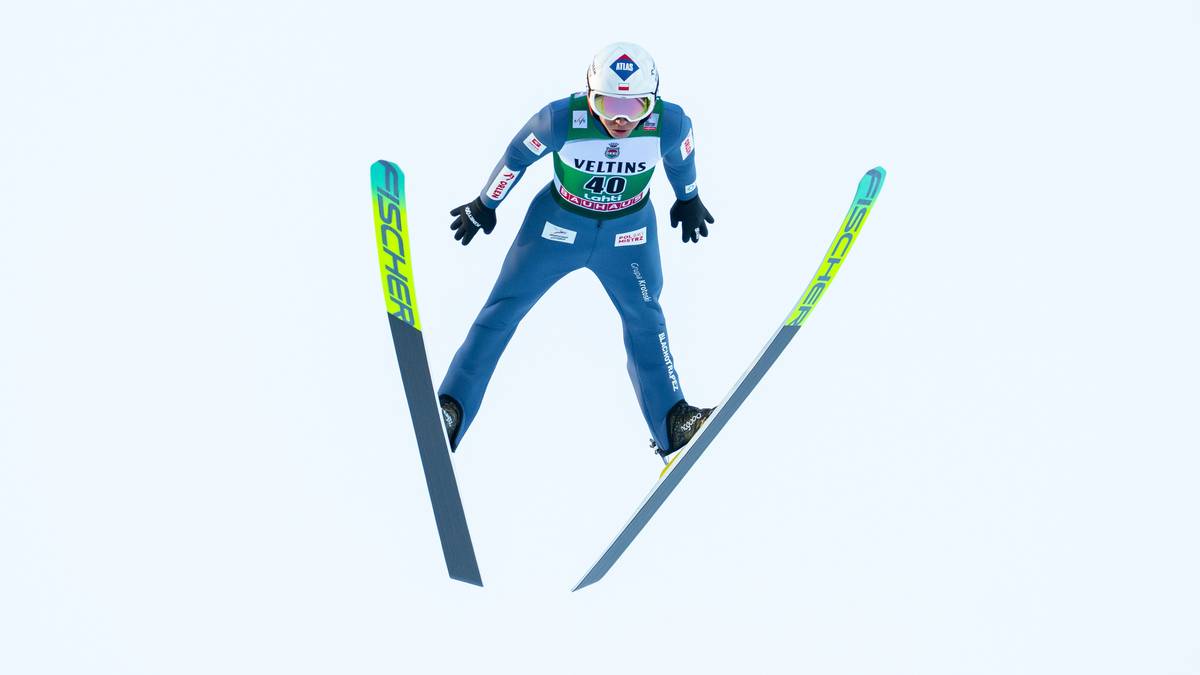 Skoczek narciarski w powietrzu podczas zawod&oacute;w, ubrany w niebieski kombinezon i biały kask, z nartami rozstawionymi w locie.