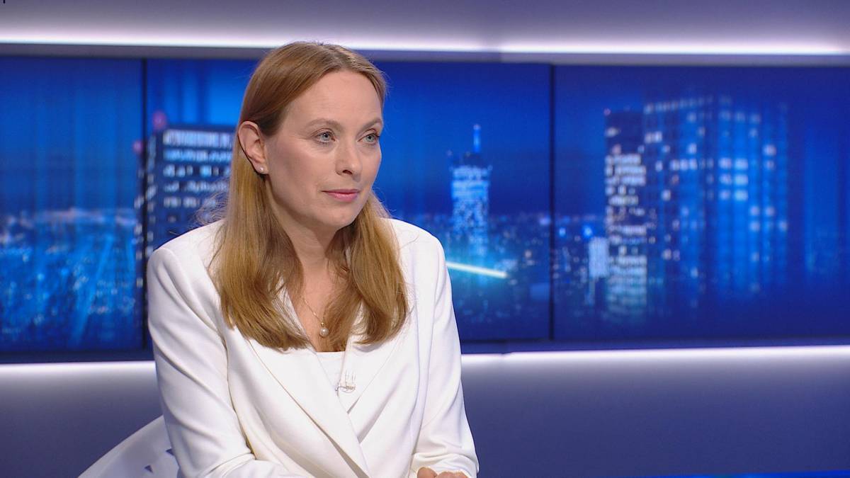 Kobieta w białej marynarce siedzi na tle ekran&oacute;w z widokiem na nocne miasto