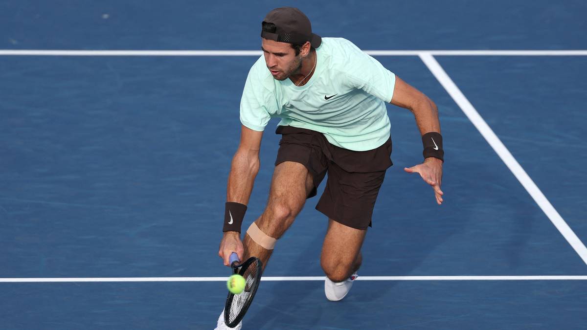 Tenisista w jasnoniebieskiej koszulce i brązowych spodenkach z rakietą tenisową i piłką na niebieskim korcie.