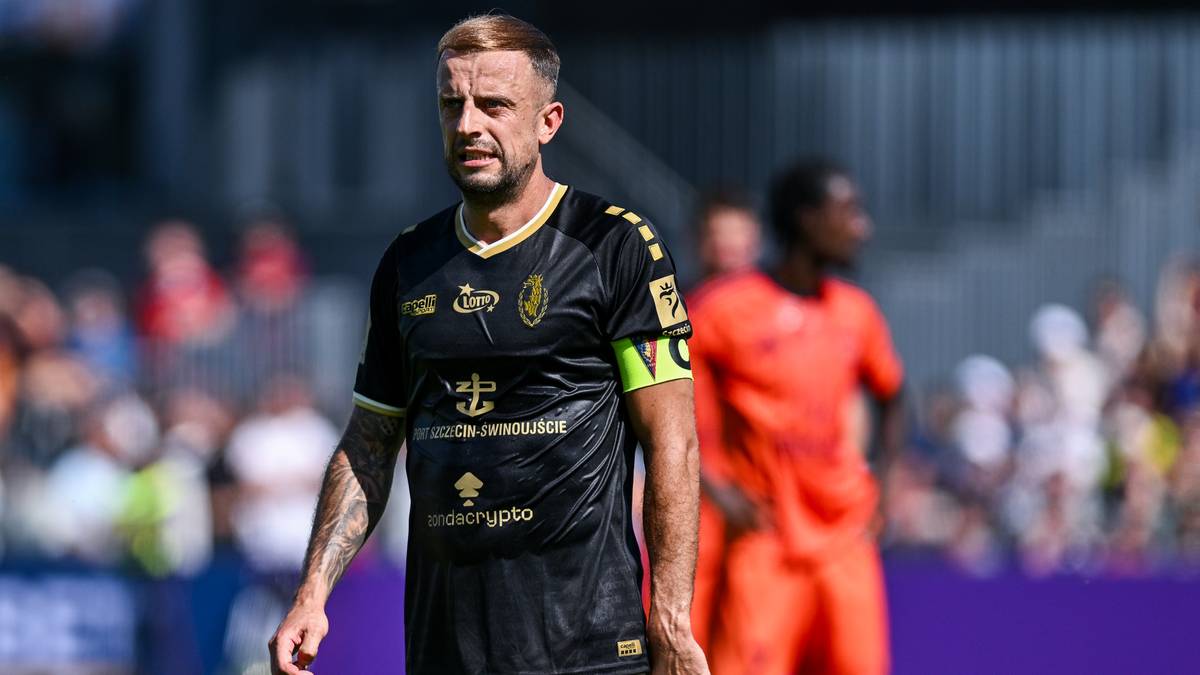 Piłkarz w czarnej koszulce z opaską na ramieniu na tle murawy i trybun.