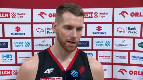 Szymon Tomczak, koszykarz, w czarnej koszulce z czerwonym dekoltem, stoi na tle ściany z logotypami sponsor&oacute;w.