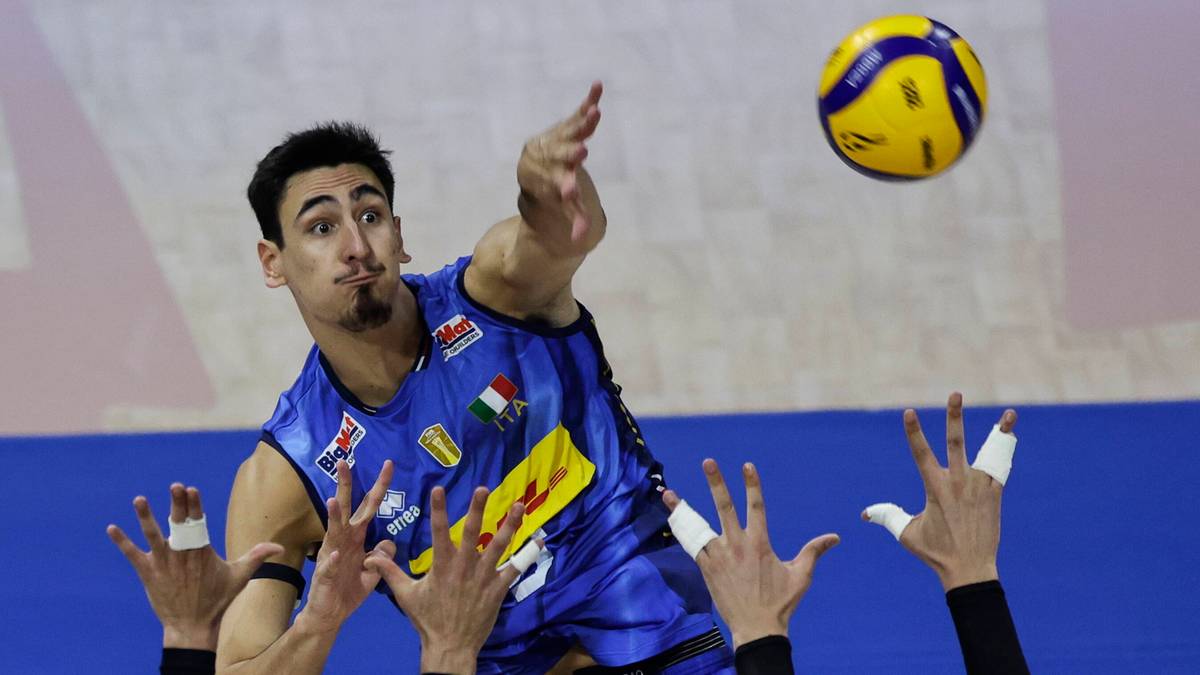 Siatkarz w niebieskiej koszulce z włoskimi barwami narodowymi wyskakuje do ataku, z piłką nad głową i dłońmi wyciągniętymi do zagrania. W tle widać ręce przeciwników próbujących zablokować piłkę.