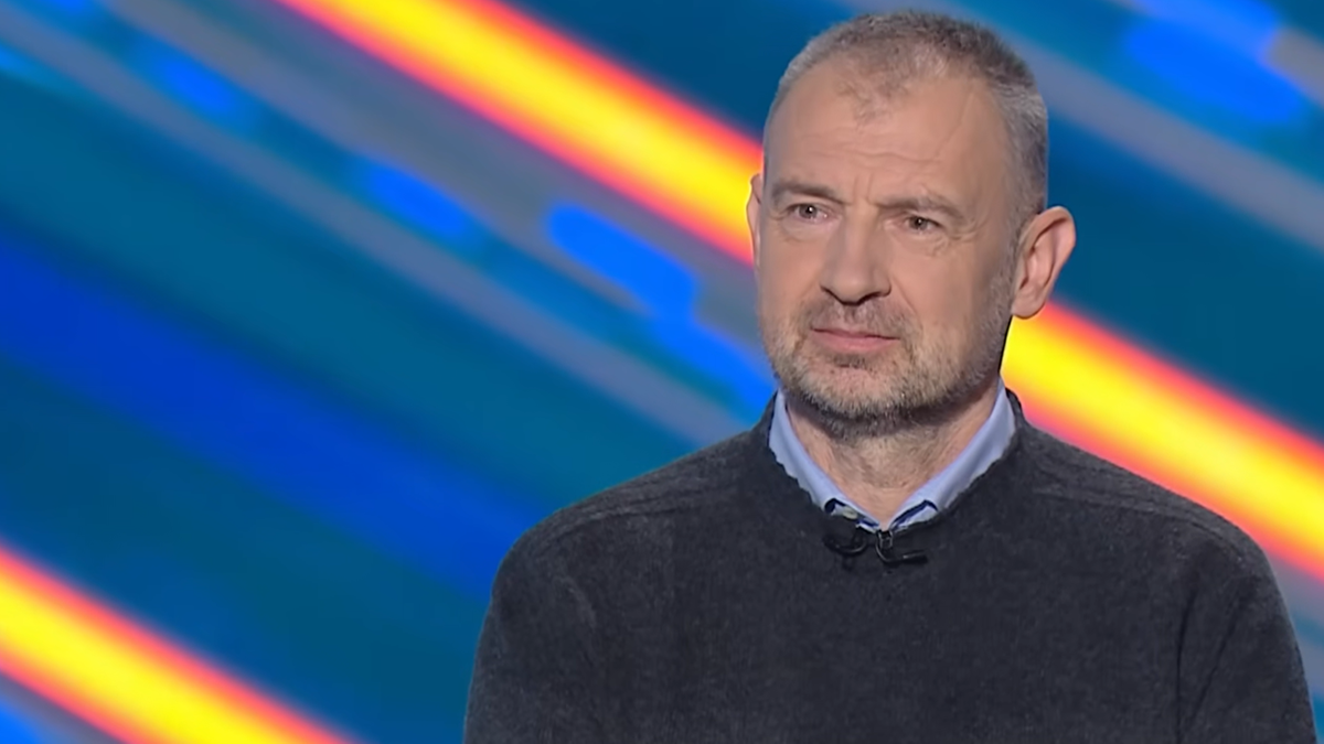 Portret mężczyzny w średnim wieku o krótkich, siwych włosach i kilkudniowym zaroście, ubranego w ciemny sweter na niebieskiej koszuli, patrzącego w kierunku obiektywu z lekko zamyślonym wyrazem twarzy. W tle widoczne są rozmyte, kolorowe pasy.