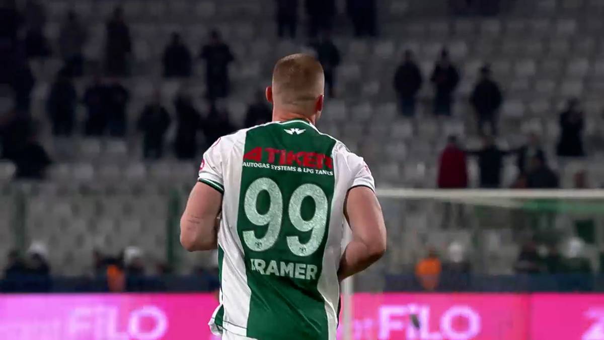 Piłkarz w białej koszulce z zielonymi wstawkami, na plecach numer 99 i nazwisko "RAMER". Zawodnik stoi tyłem do widza, na tle trybun wypełnionych widzami.