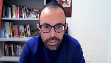 Mężczyzna w okularach i niebieskiej bluzie z brodą siedzi przed regałami pełnymi książek.