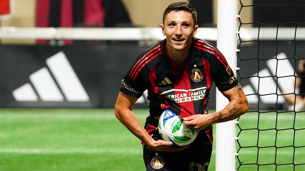 Piłkarz w czarno-czerwonej koszulce z piłką pod pachą, uśmiechnięty, stoi przy bramce.