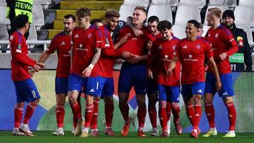 Drużyna piłkarska w czerwonych strojach świętuje gola, wielu zawodnik&oacute;w przybija sobie piątki i obejmuje się.