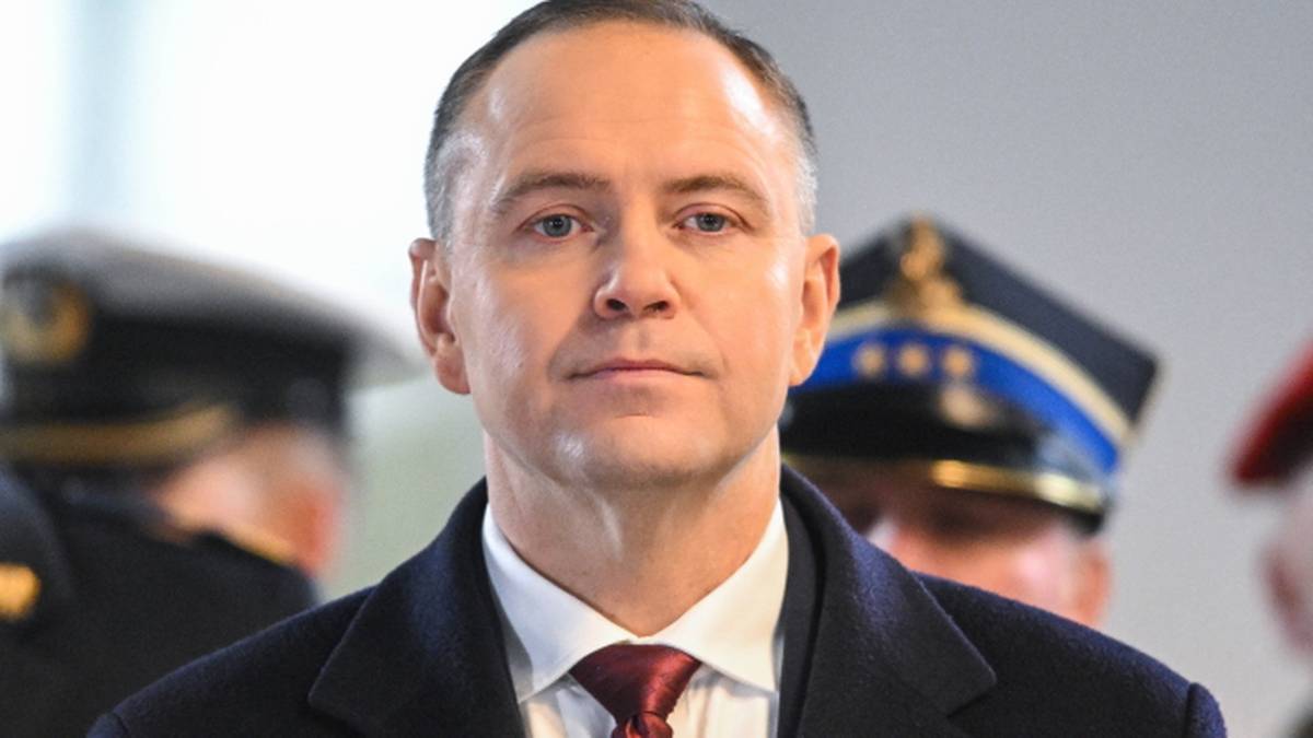 Prezydent Rzeczypospolitej Polskiej Karol Nawrocki w garniturze i krawacie, w tle widoczni inni mężczyźni w mundurach.