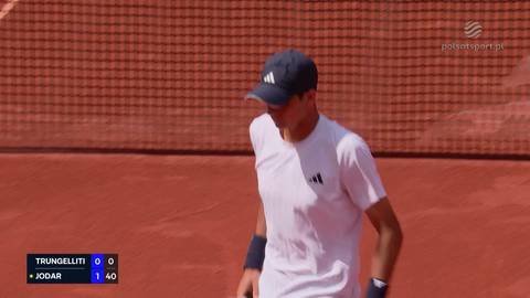 Tenisista w białej koszulce i czapce z daszkiem, z opaskami na nadgarstkach, stoi na korcie tenisowym.