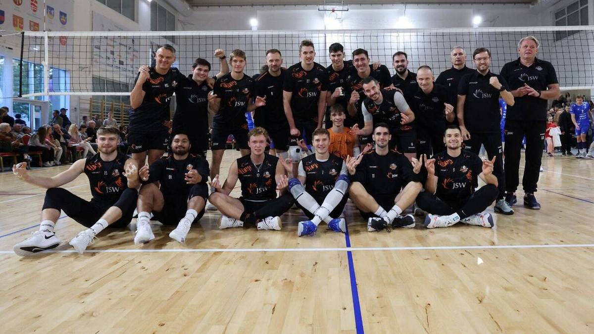 Drużyna siatkówki Jastrzębski Węgiel pozuje do wspólnego zdjęcia po wygranym turnieju. Zawodnicy ubrani są w stroje sportowe, niektórzy trzymają puchary. W tle siatka do gry i hala sportowa.