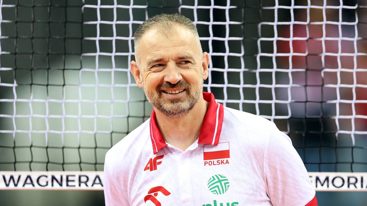 Uśmiechnięty mężczyzna w białej koszulce z czerwonym kołnierzykiem i polską flagą na piersi, stoi na tle siatki.