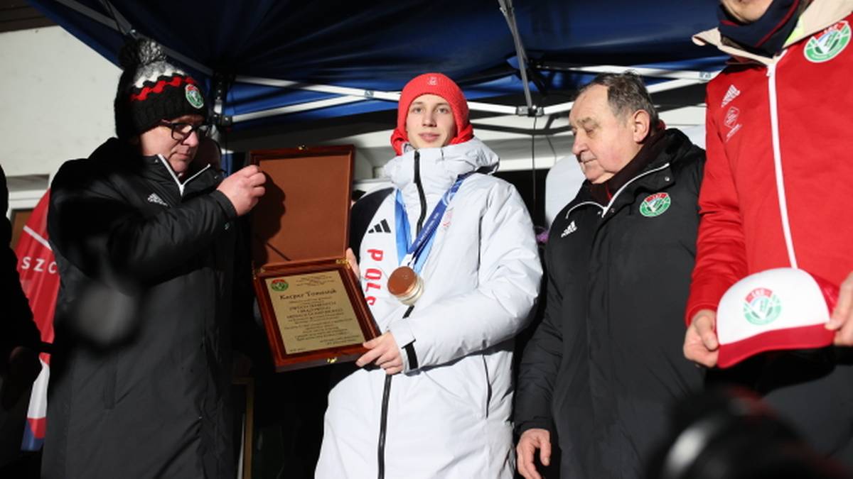 Młody skoczek narciarski Kacper Tomasiak w białej kurtce i czerwonej czapce odbiera medal i dyplom na podium, otoczony przez mężczyzn.