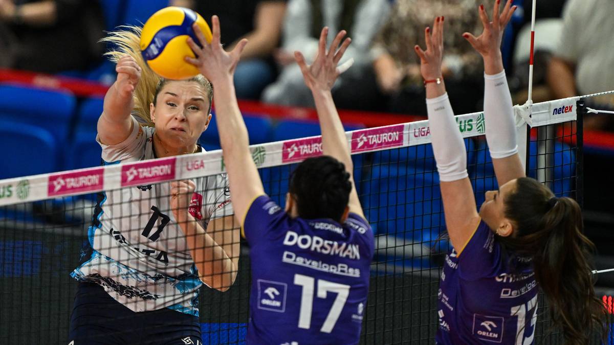 Siatkarka w biało-niebieskim stroju z numerem 11 na plecach atakuje piłkę nad siatką, podczas gdy dwie zawodniczki w fioletowych strojach pr&oacute;bują ją zablokować.
