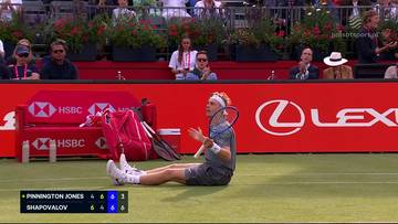 Tenisista Denis Shapovalov siedzi na trawiastym korcie, trzymając rakietę, z wyrazem satysfakcji na twarzy po udanej akcji. W tle widać widz&oacute;w i logotypy sponsor&oacute;w.