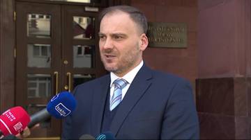 Mężczyzna w garniturze i krawacie przemawia do mikrofon&oacute;w.