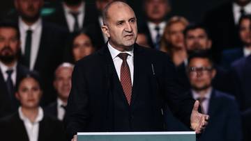 Polityk w garniturze przemawia za m&oacute;wnicą, otoczony przez innych ludzi.