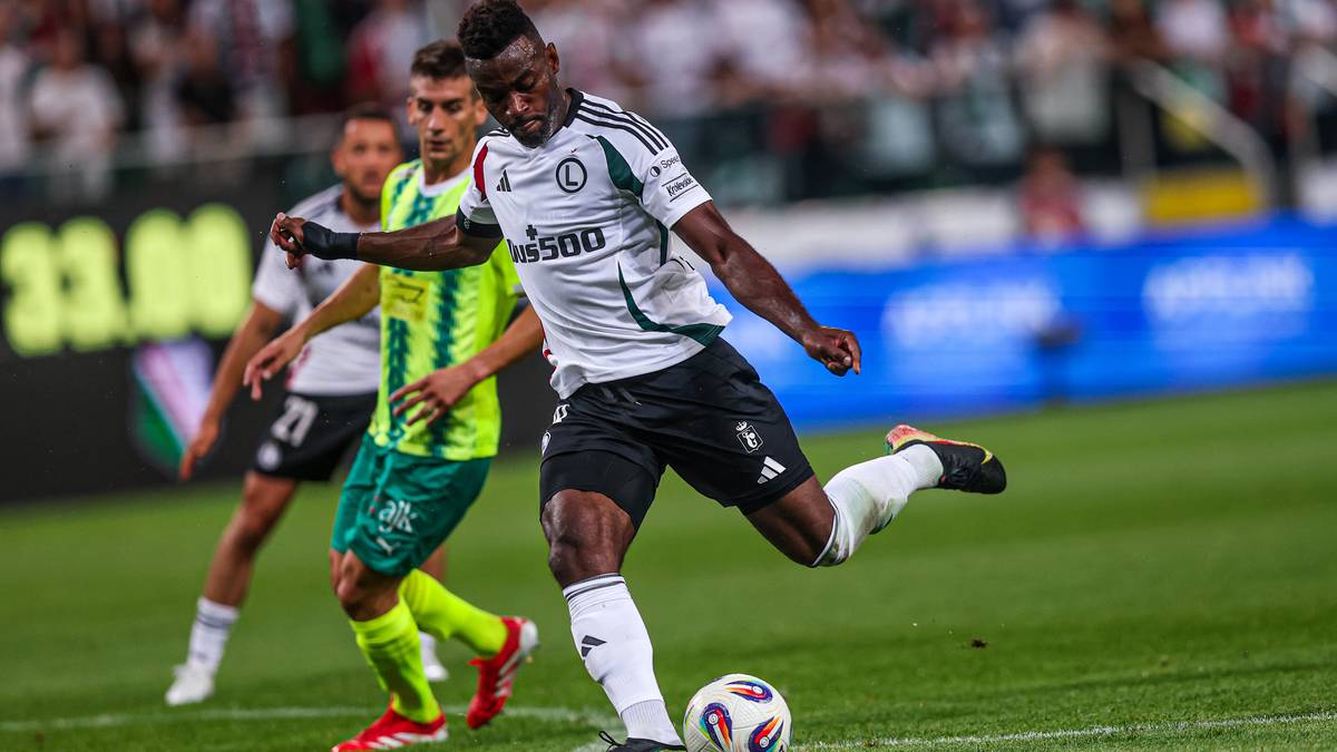 Piłkarz w białej koszulce Legii Warszawa kopie piłkę na zielonej murawie, ścigany przez przeciwnika w ż&oacute;łtej koszulce.