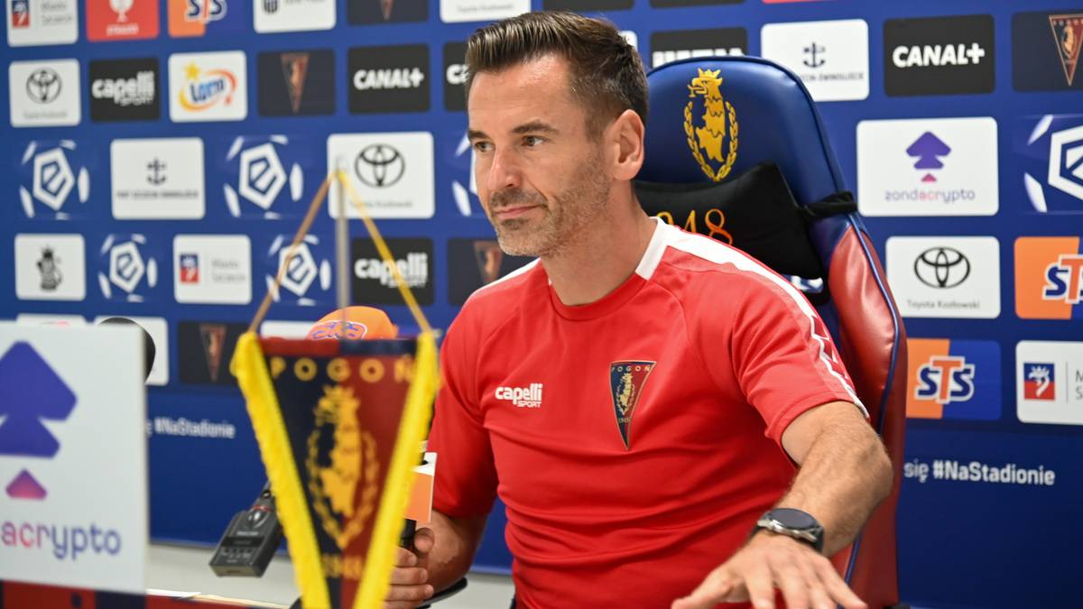 Mężczyzna w czerwonej koszulce z symbolem klubu siedzi na krześle, trzymając w ręce flagę z barwami klubowymi. W tle widać ściany ozdobione logotypami sponsorów i nazwami kanałów.