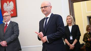 Polityk w garniturze i okularach idzie korytarzem, wykonując gest pokazujący kciuk w g&oacute;rę.