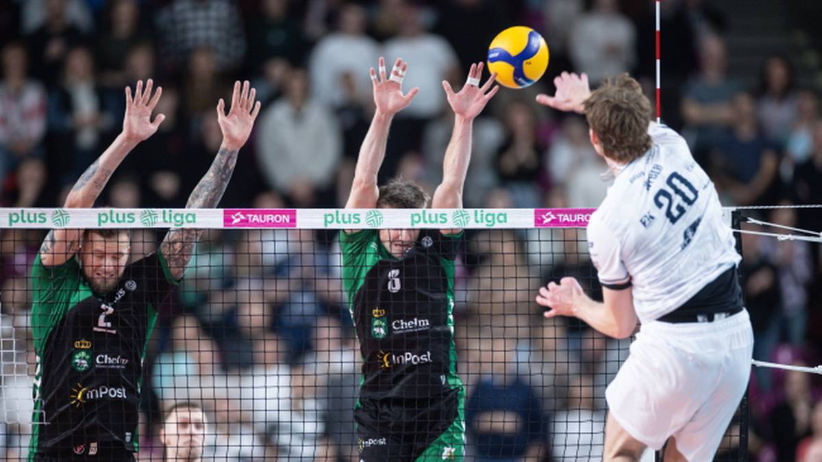 Siatkarz w białej koszulce z numerem 20 wykonuje atak nad siatką, próbując ominąć blokujących przeciwników w zielonych strojach.