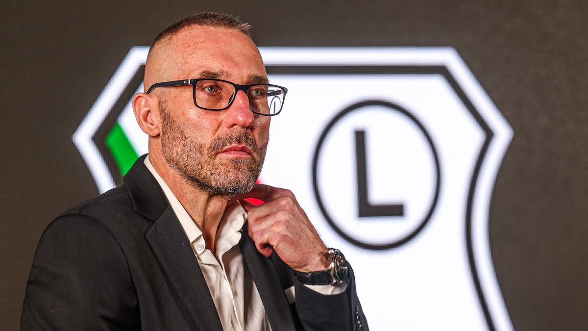 Mężczyzna w okularach na tle logo Legii Warszawa.