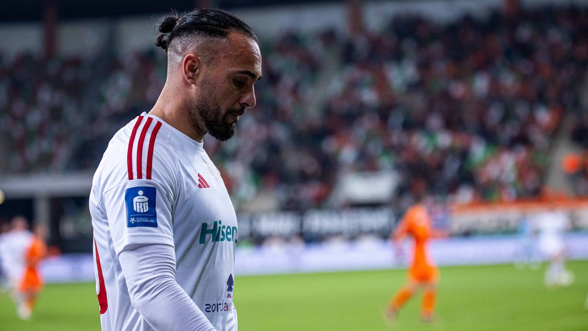 Piłkarz w białej koszulce z czerwonymi pasami na ramieniu patrzy w bok na boisku piłkarskim.