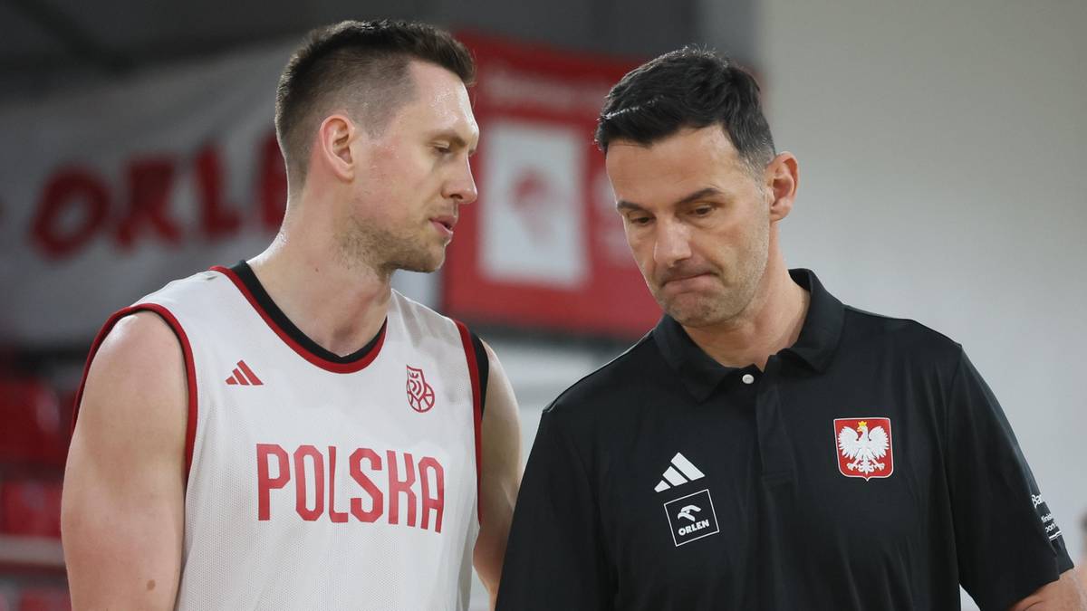 Dw&oacute;ch mężczyzn w strojach sportowych, jeden w białej koszulce z napisem "POLSKA" i czerwonymi spodenkami, drugi w czarnej koszulce z logo "Adidas" i orłem. Mężczyzna w czarnej koszulce trzyma tablicę taktyczną. W tle widać fragmenty trybun, baner z napisem "ORLEN" oraz innego mężczyznę w stroju sportowym z piłką do koszyk&oacute;wki.