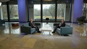 Dw&oacute;ch mężczyzn w garniturach siedzi naprzeciwko siebie na kanapach w nowoczesnym wnętrzu, przed stolikiem kawowym. W tle duże okna z widokiem na park.