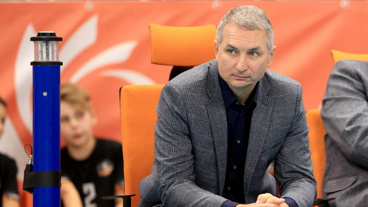 Mężczyzna w szarym garniturze siedzi na krześle, z rękami złożonymi na kolanach. W tle widać chłopca.