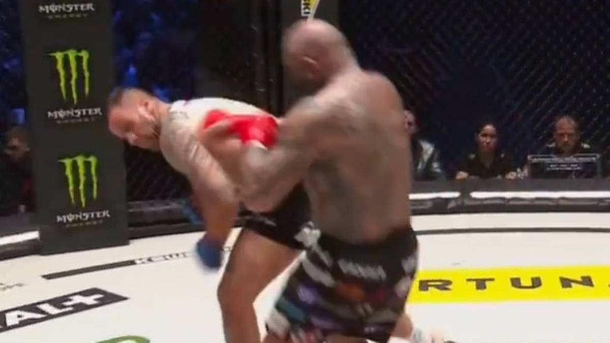 Dw&oacute;ch zawodnik&oacute;w sport&oacute;w walki w oktagonie. Jeden zawodnik w białej koszulce i czarnych spodenkach jest powalany ciosem przez przeciwnika w kolorowych spodenkach.