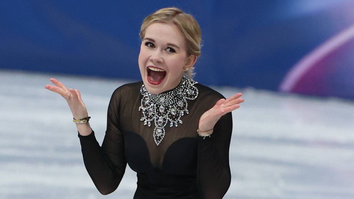 Łyżwiarka figurowa w czarnej sukience z ozdobnym kołnierzem stoi na lodowisku z uniesionymi rękami, patrząc w stronę widza z ekspresją zaskoczenia lub radości. W tle widoczny jest niebieski baner z napisem "MILANO CORTINA 2026".