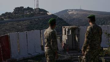 Dw&oacute;ch żołnierzy w mundurach wojskowych i zielonych beretach stoi odwr&oacute;conych plecami do widza, patrząc na g&oacute;rzysty krajobraz z wieżą telekomunikacyjną na wzg&oacute;rzu.