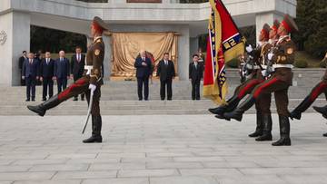 Żołnierze w historycznych mundurach defilują przed monumentem, niosąc flagi. W tle grupa mężczyzn obserwuje ceremonię.