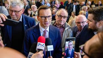 Mateusz Morawiecki w okularach i garniturze odpowiada na pytania dziennikarzy trzymających mikrofony.