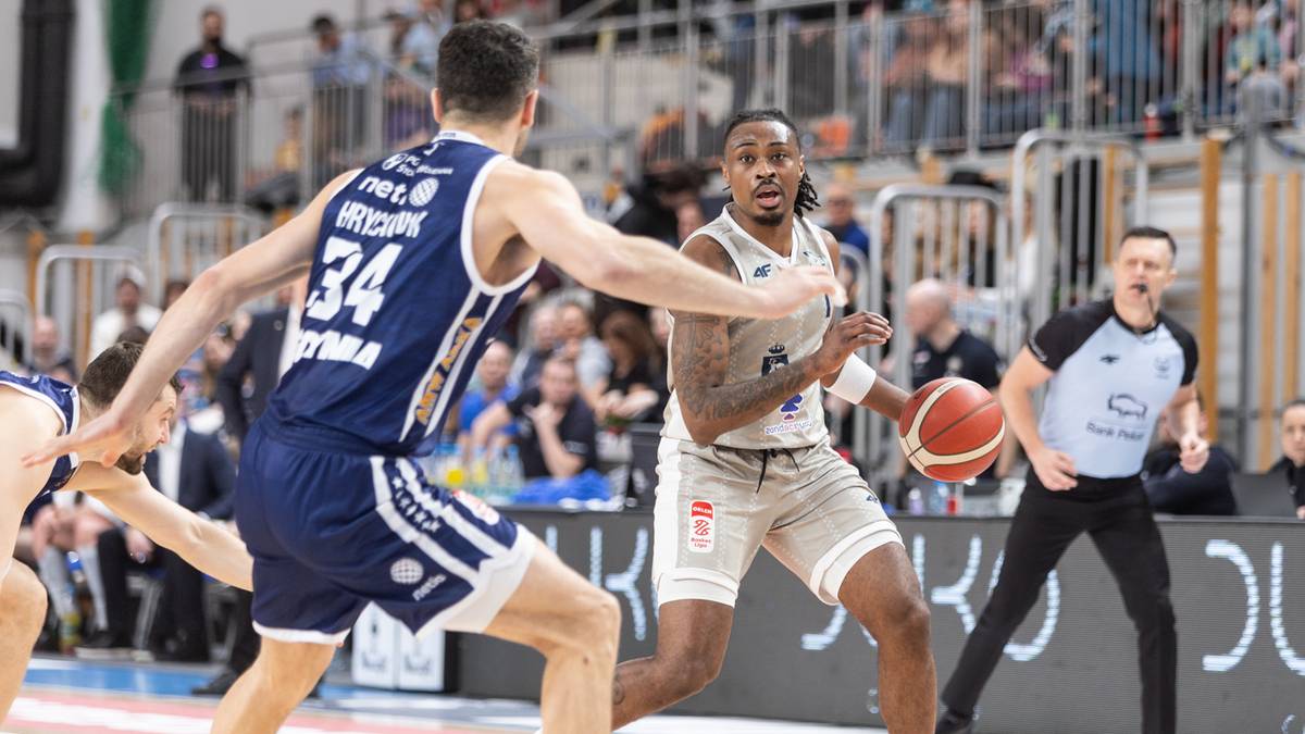 Dw&oacute;ch koszykarzy w akcji, jeden z piłką, drugi broniący, z arbitrem w tle.