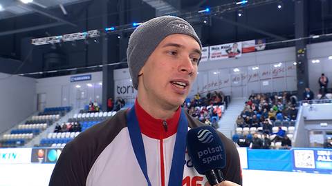 Marek Kania w czapce i stroju sportowym z medalem na szyi udziela wywiadu z mikrofonem Polsat Sport na tle trybun lodowiska.