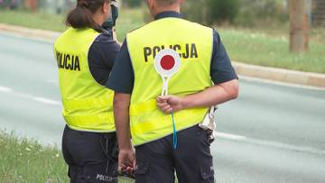 Dwóch policjantów w kamizelkach odblaskowych z napisem POLICJA, jeden z nich trzyma znak "STOP".