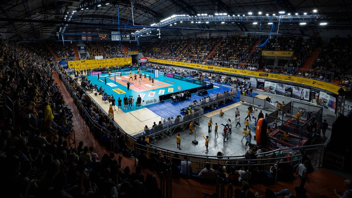 Boisko do siatkówki z zawodnikami i widownią podczas meczu w dużej hali sportowej.